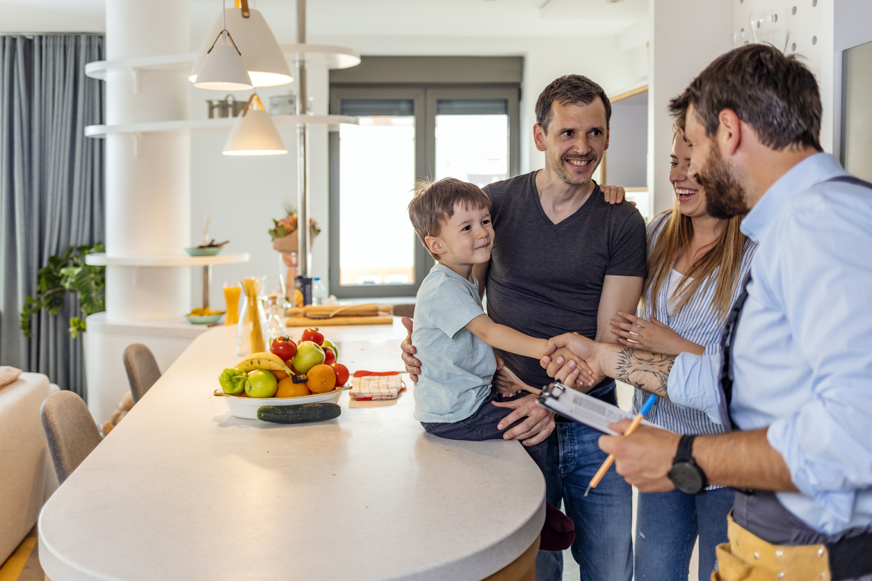 Professional plumber in uniform talking to mother and father clients and shaking hands with their small son indoors.